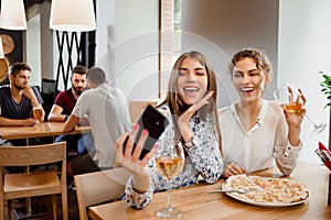 Two positive girls smiling and making selfie with wine in restaurant.