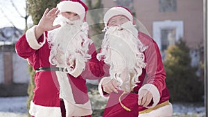 Two positive men in red Santa costumes waving and opening red gifts sack. Portrait of joyful smiling Santas bringing