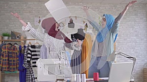 Two positive young muslim women fashion designers in national headscarves throw papers for joy slow mo