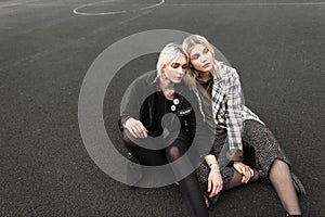 Two pretty beautiful sisters in trendy casual youth clothes resting on basketball court outdoors. Cute blonde girlfriends in