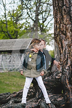 Two pretty cute children boy and girl standing near big tree trunk in summer park outdoors