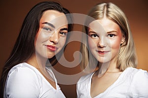 two pretty diverse girls happy posing together: blond and brunette, caucasian and asian on brown background, lifestyle