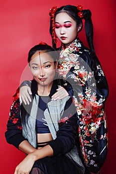 Two pretty geisha girls friends: modern asian woman and traditional wearing kimono posing cheerful on red background