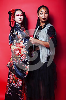 Two pretty geisha girls friends: modern asian woman and traditional wearing kimono posing cheerful on red background