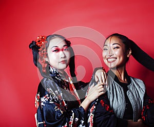 Two pretty geisha girls friends: modern asian woman and traditional wearing kimono posing cheerful on red background