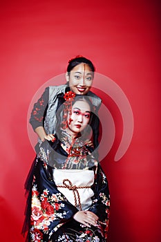 Two pretty geisha girls friends: modern asian woman and traditional wearing kimono posing cheerful on red background