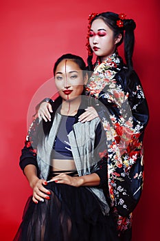 Two pretty geisha girls friends: modern asian woman and traditional wearing kimono posing cheerful on red background