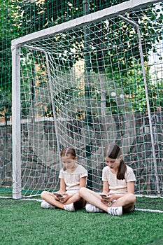 two pretty girls in identical clothes using phone on sports ground or athletic field. kids are surfing in internet