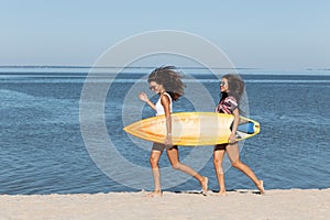 Two pretty girls are walking along the sandy beach near the sea and holding together a yellow surfboard in their hands