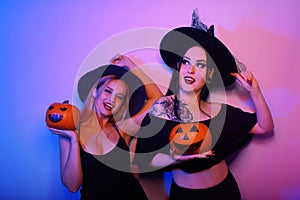 Two pretty witch poses with pumpkins for a Halloween photo. Girls in witch hats in neon dance at the party