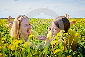 Two pretty women on a field with green grass and yellow dandelion flowers in a sunny day with blue sky. Girls having rest and fun