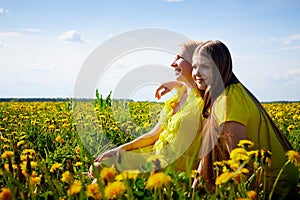 Two pretty women on a field with green grass and yellow dandelion flowers in a sunny day with blue sky. Girls having rest and fun