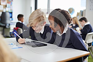 Two primary school kids sitting together at desk in a classroom using a tablet computer and stylus