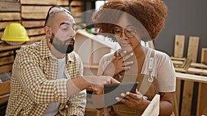 Two professional carpenters speaking business at carpenter workshop, expertly navigate touchpad amidst timber and furniture