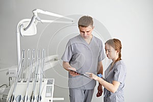 Two professional confident dentists posing near dental chair at clinic. Dentist and assistant in exam room smiling