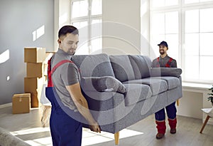 Two professional relocation service workers in overalls move sofa in customer& x27;s apartment.
