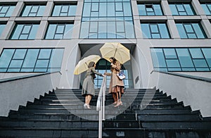 Two stylish businesswomen are exiting an office building, holding yellow umbrellas on a cloudy day, representing