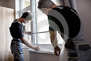 Two professional workers in uniform using tape measure while measuring window and making notes for installing blinds