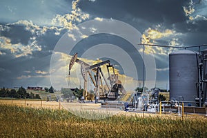 Two pump jacks and tanks and oil field equipment in fenced area in pasture under dramatic skies with houses on hill on horizon -