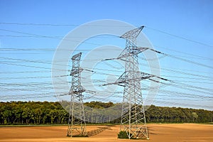 Two pylons of high voltage power lines located on a plowed field, against a background of blue sky and green forest. Shadows from
