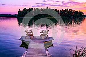 Two rattan chairs and glasses of red wine on a pier overlooking a lake at sunset in Finland