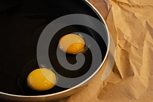 Two raw eggs with bright yellow yolks and transparent whites with a clots in a black frying pan on paper. The concept of  cooking