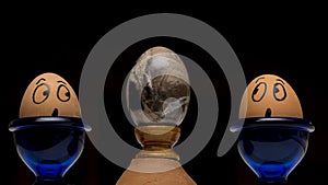 Two real hand-painted eggs look in amazement at a stone egg of volcanic origin against a black background