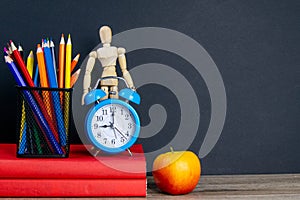 Two red books stand on the table, on the books there are blue watches and a glass with multi-colored pencils, behind the clock