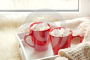 Two red cup of hot chocolate with marshmallow on white windowsill with furskin for rest. Holiday.