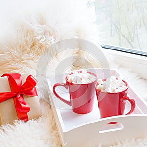 Two red cup of hot chocolate with marshmallow on white windowsill with furskin for rest. Holiday.