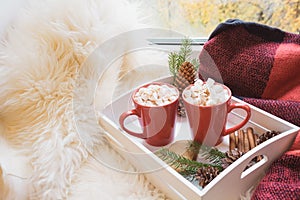 Two red cup of hot chocolate with marshmallow on white windowsill with furskin for rest. Holiday. Christmas morning.