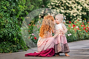 Two red-haired sisters in long linen dresses are resting on the lake in the park The older girl hugs and kisses the baby
