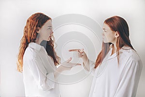 Two red-haired sisters stand isolated on a white background in spacious oversized shirts. TWO young girls accuse each