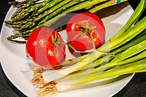 Two, red, ripe tomatoes, six green and white scallions with a group of asparagus, on white plate