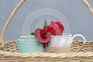 Two red roses on couple cups of tea or coffee in a wooden rattan basket on blue background. Relationship and valentine day concept