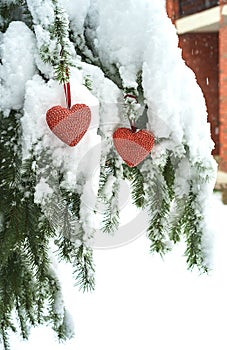 Two red textile hearts hanging on heavy snowy fir branch, near red brick house. Merry Christmas, Happy New Year and Valentine`s