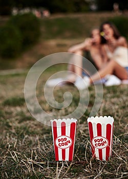 Two red and white boxes of popcorn, standing on green pale grass in city park, with blurred people at the background, Close-up