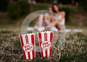 Two red and white boxes of popcorn, standing on green pale grass in city park, with blurred people at the background, Close-up
