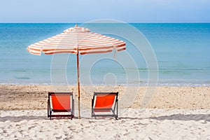 Two red wooden chairs and beach umbrella setting on white sand with seascape and blue sky in the background.