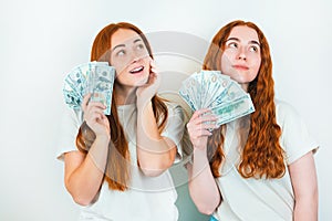 Two redheaded young women both holding cash like playing cards looking thoughful standing on isolated white backgroung, money