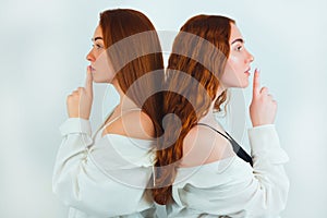 Two redheaded young women both holding fingers near lips showing keep silence sign standing with their backs turned on