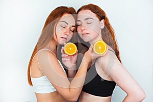 Two redheaded young women both holding half of orange in their hands stand facing each other on isolated white background, beauty