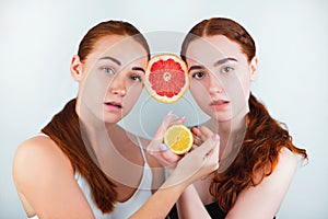 Two redheaded young women clamping half of grapefruit between their foreheads holding half of orange in hands standing on isolated