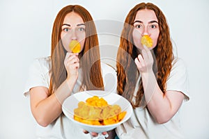 Two redheaded young women holding crispy potatoe chips near their mouths standing on isolated white backgroung, junk food concept