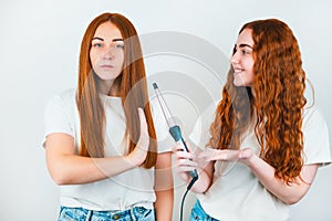 Two redheaded young women standing on isolated white backgroung, one wants to curl her friend`s hair with styler, but she refuses