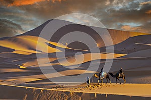 Two Riders And Their Handler Travel Through The Saharan Desert On Their Camels In Morocco