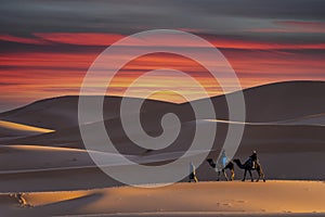 Two Riders And Their Handler Travel Through The Saharan Desert On Their Camels In Morocco