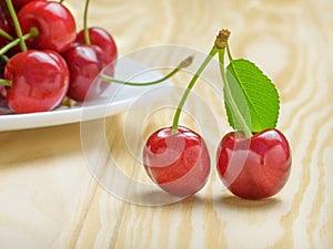 Two ripe red cherries with a green leaf next to a white plate filled with cherries. Light wood background
