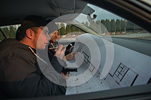 Two robbers sitting inside a car with guns looking at the blueprint of the building