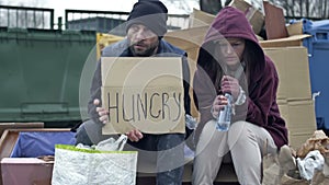 Two sad homeless men sit by the rubbish bins. Man and a woman are poor and dirty dressed. The man is holding HUNGRY